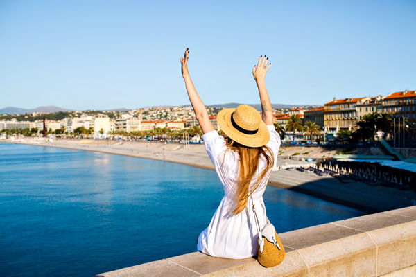 In the photo, a tourist in a white dress and a straw hat on the seashore with her hands raised up